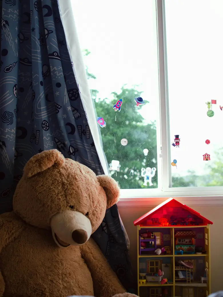 Teddybär sitzt am Fenster eines Kinderzimmers, auf der Scheibe kleben Raketen- und Roboteraufkleber